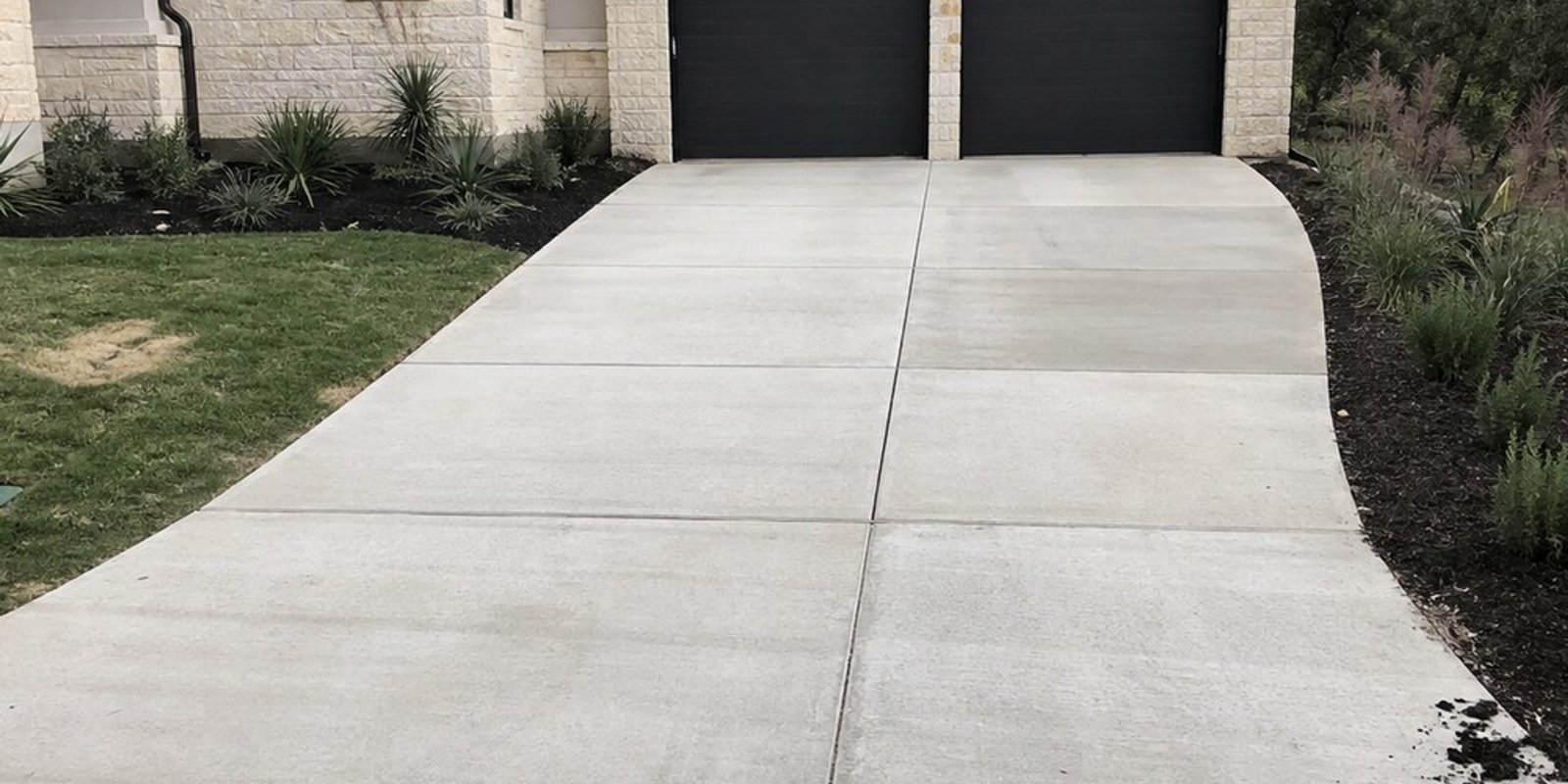 Broom-finish concrete driveway with saw-cut control joints leading to a two-car garage with dark doors on a limestone home with a metal roof.