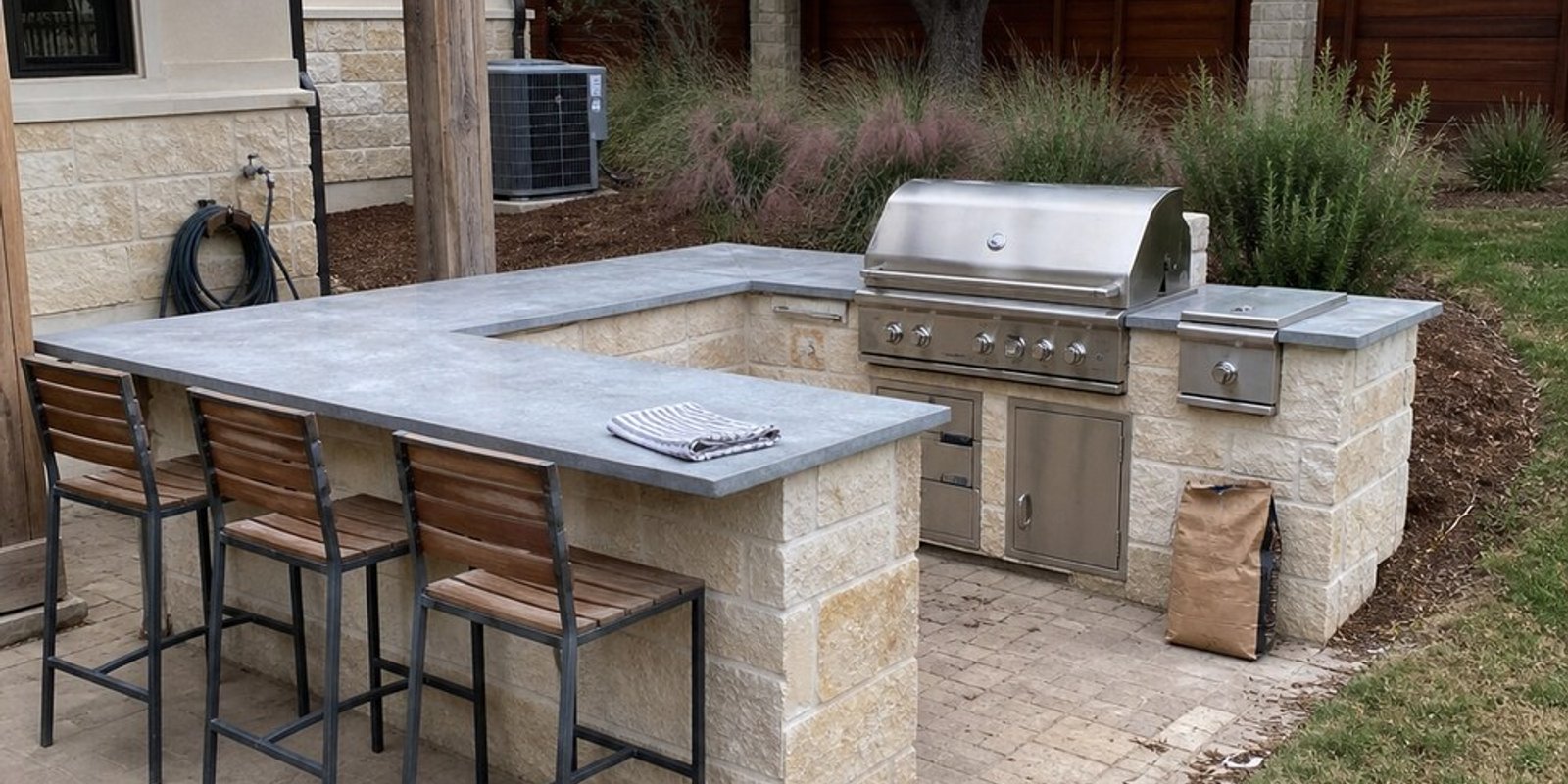 U-shaped outdoor kitchen with a cut limestone block base, gray stone countertop with bar stools, and a built-in stainless steel grill and side burner on a paver patio.