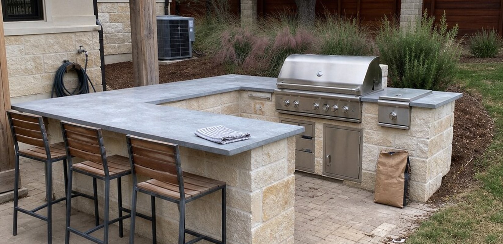 U-shaped outdoor kitchen with a cut limestone block base, gray stone countertop with bar stools, and a built-in stainless steel grill and side burner on a paver patio.